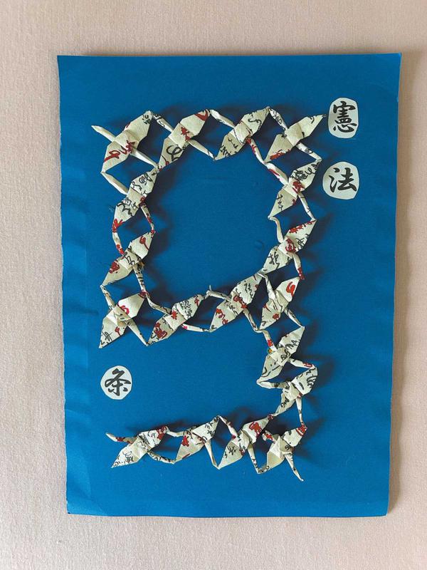 A blue foam board carries the Japanese word "Constitution" at its upper right corner. At the center, 18 hand-folded paper cranes are arranged to form the number "9" — symbolizing Article 9 of the Constitution of Japan, the peace clause. JIANG XUEQING/CHINA DAILY