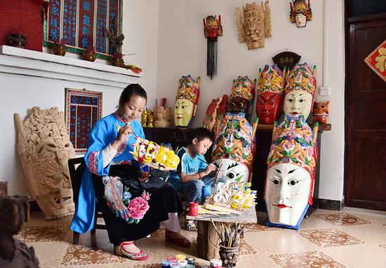 A young woman and a child paint wood masks for Dixi Opera performances. (XU LIN/CHINA DAILY/YUAN QINSHU/FOR CHINA DAILY)