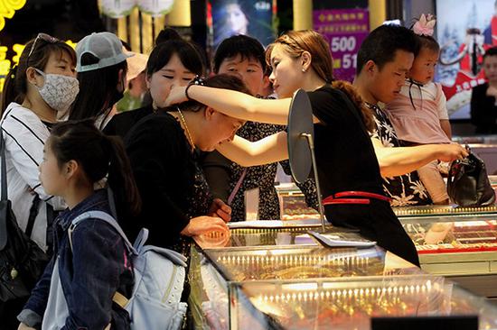 A saleswoman helps a customer try on a gold necklace at a jewelry store in Xuzhou, Jiangsu province. Gold prices may stay high due to economic uncertainty (which pushes investors to perceived safe havens) and jewelry demand, analysts said.  (Photo by Yu Tian/for China Daily)