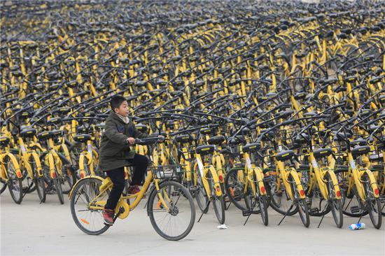 An Ofo customer rides past piles of the company's bikes in Xiangyang, Hubei province. (Photo by Guo Qi/For China Daily)