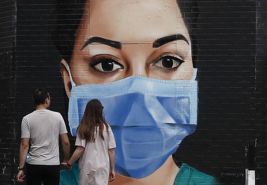 A couple walk hand in hand past a mural of a masked NHS (National Health Service) worker painted on a wall in London, Britain, on April 26, 2020. (Xinhua/Han Yan)
