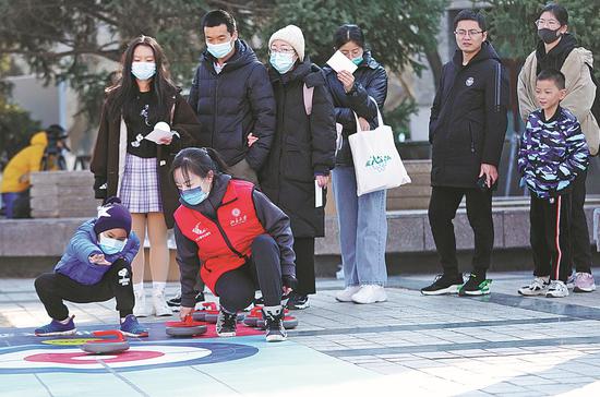 A volunteer who will serve at the Beijing 2022 Winter Olympics helps people experience floor curling in Beijing on Sunday at an event organized by Peking University to celebrate the 36th International Volunteer Day and pass on the spirits of volunteerism. (Photo by Fang Fei/for China Daily)