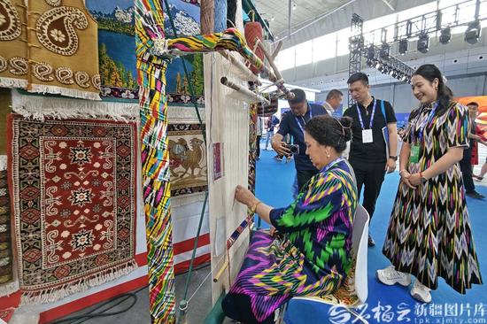 6月25日，在非物质文化遗产展位上，帕米尔·阿力甫（左）在为参展商介绍织毯产品。